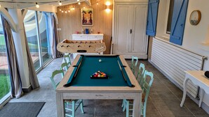 Una mesa de billar con superficie de fieltro verde, rodeada de sillas verde claro, en una habitación con paneles de madera y un armario blanco.