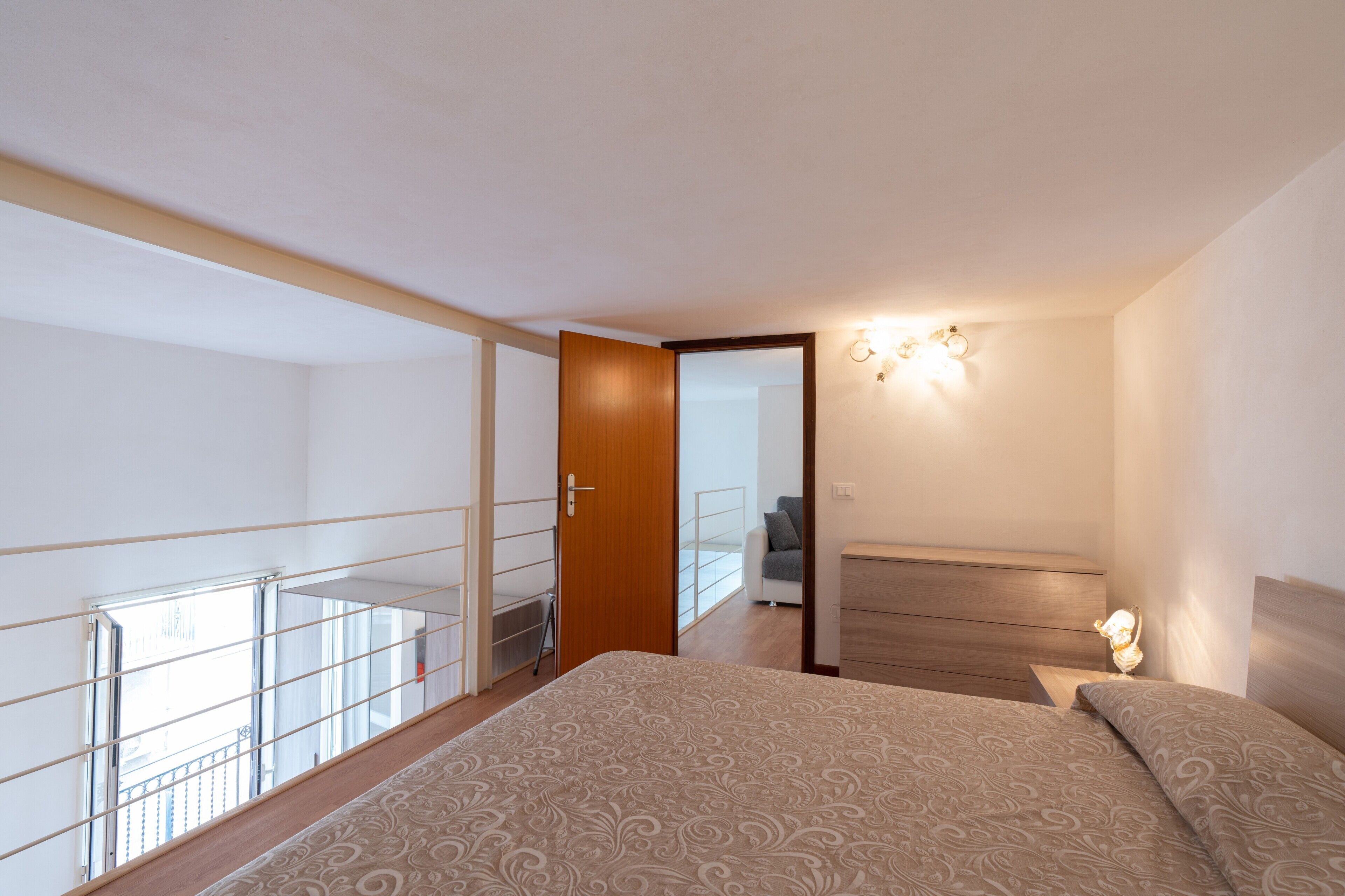 Ein Schlafzimmer mit Bett, Nachttisch, Wandleuchte, einem Balkon mit Geländer und einer Tür zu einem anderen Raum.