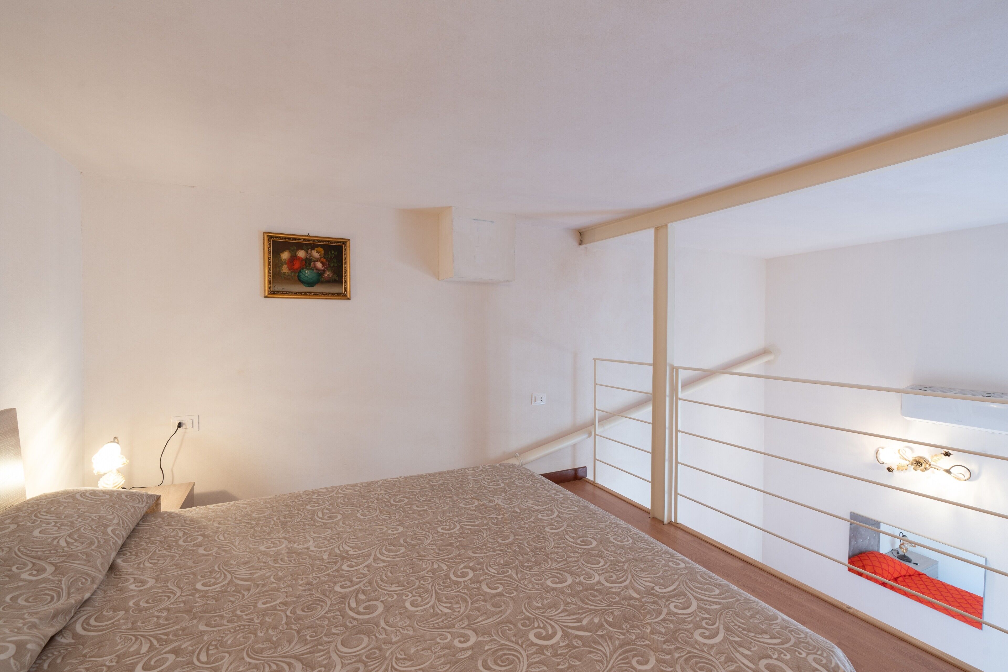 Une chambre à coucher avec un lit, une table de chevet, une lampe fixée au mur, un tableau, un balcon avec une balustrade et une fenêtre.