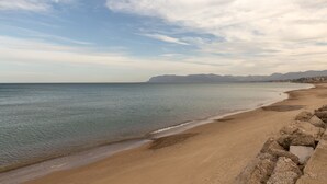 Una playa con costa rocosa, agua tranquila y montañas distantes bajo un cielo parcialmente nublado.