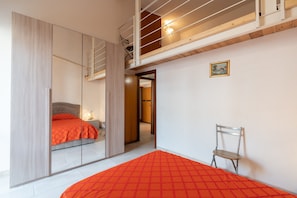 Un dormitorio con cama, una silla, un espejo y una puerta que da a otra habitación.