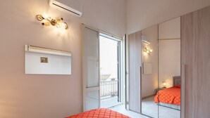 Un dormitorio con cama, una puerta corrediza a un balcón, aire acondicionado montado en la pared y una mesita de noche con lámpara.
