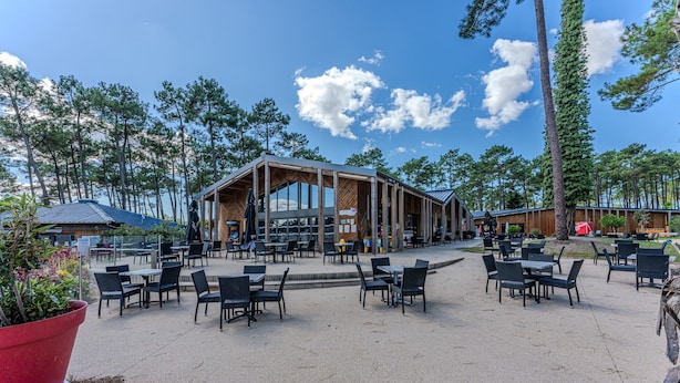 Een terras met tafels en stoelen, een gebouw met grote ramen, en een bebost achtergrond.
