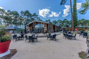 Un espace extérieur aménagé pour les repas, avec des tables et des chaises, un bâtiment doté de grandes fenêtres et un paysage boisé en arrière-plan.