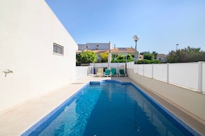 Une piscine avec un bord carrelé, un mur blanc percé d’une fenêtre et une clôture.