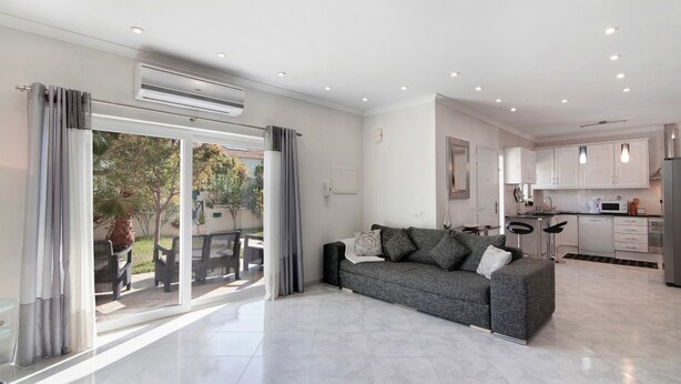A modern living room with a grey sofa, a dining area with chairs, a kitchen with white cabinets, and a sliding glass door leading to an outdoor area. - Villa 'Alonzo' with Private Pool, Wi-Fi and Air Conditioning (Albufeira)