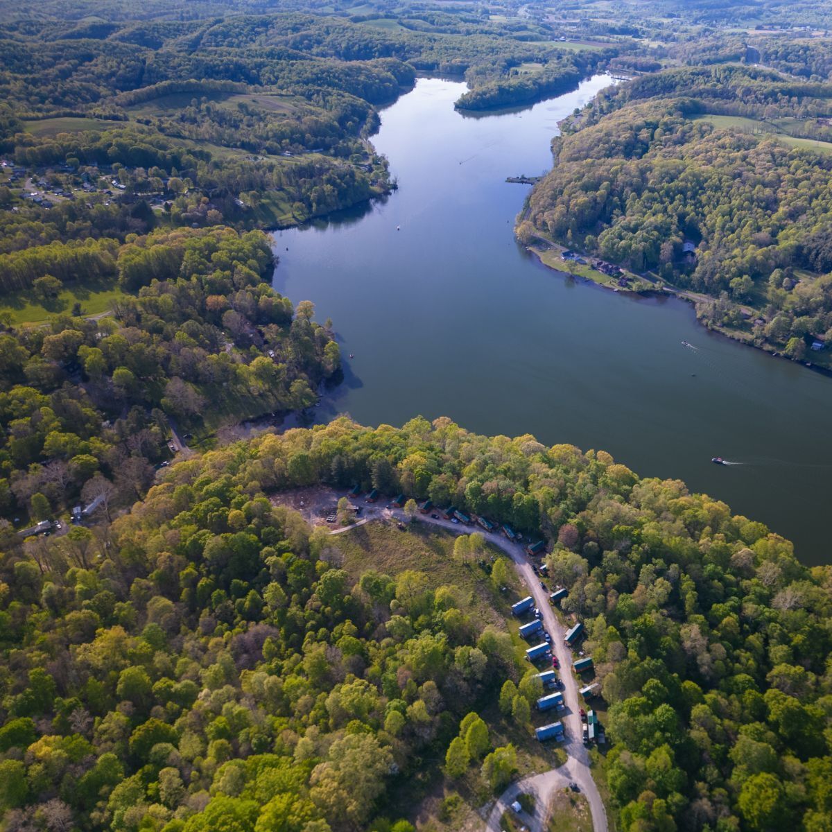 An aerial view of a lake surrounded by dense forest, with a row of cabins along the shoreline.