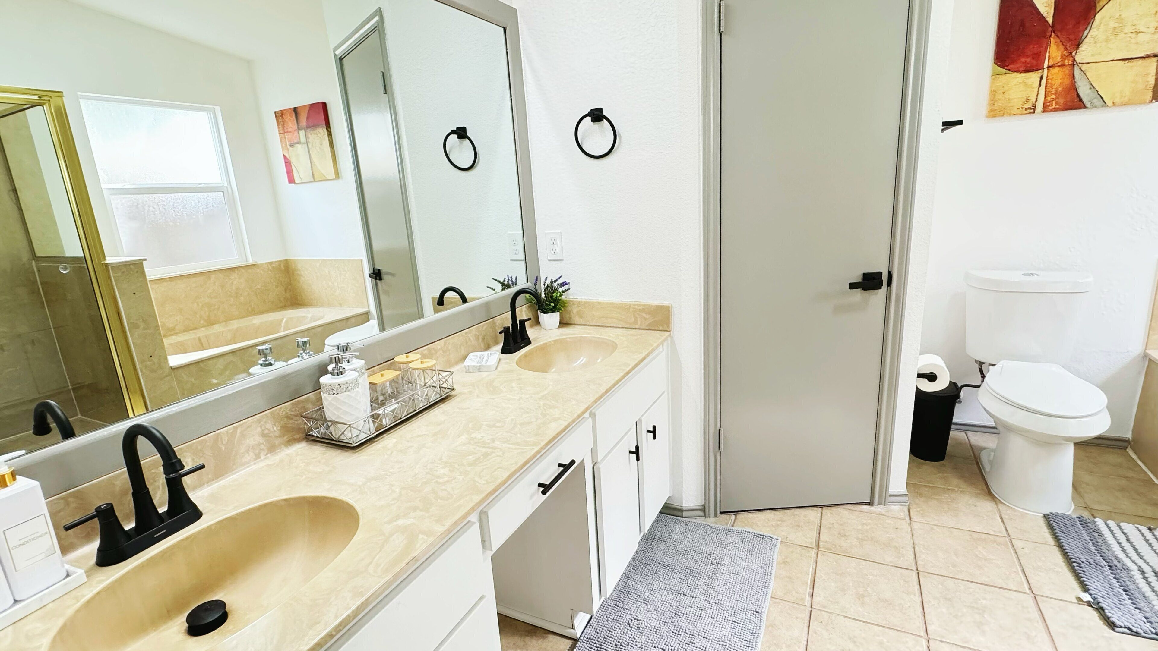 A bathroom with a beige countertop, a large mirror, a black faucet, a white toilet, and a trash bin.