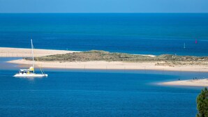 Un paysage côtier avec un voilier sur une eau calme, une plage de sable et une petite île couverte de végétation.