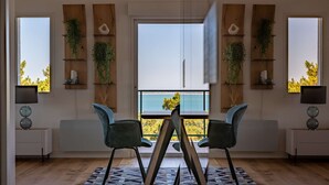 Un espace repas moderne avec vue sur la mer, comprenant une table avec des chaises, des étagères en bois ornées de plantes et un tapis à motifs.