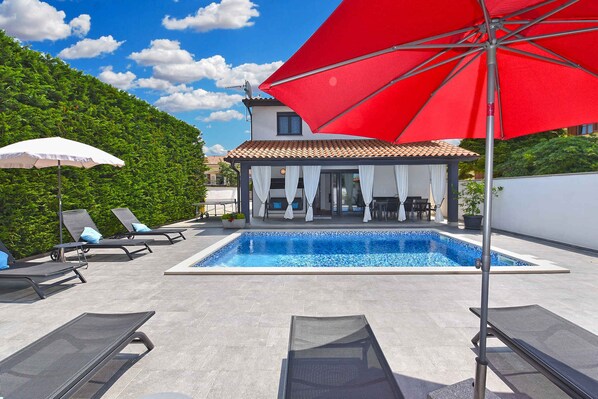 A swimming pool with lounge chairs and umbrellas, a covered patio area, and a building with a tiled roof.