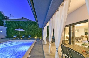 A modern outdoor pool area with a glass-enclosed dining area and a green hedge wall.