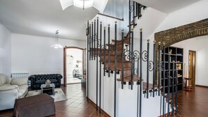 Un vaste salon comprenant un canapé en cuir noir, un fauteuil beige et un pouf marron. Un escalier en bois, avec des balustrades noires, mène à l’étage supérieur.