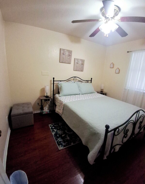 A bedroom with a bed, a nightstand, a lamp, a small table, a chair, a ceiling fan, and a window with curtains.