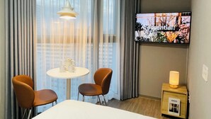 Habitación de hotel con televisión en la pared, una mesa redonda con sillas, una lámpara en una mesita de noche y una ventana con cortinas.