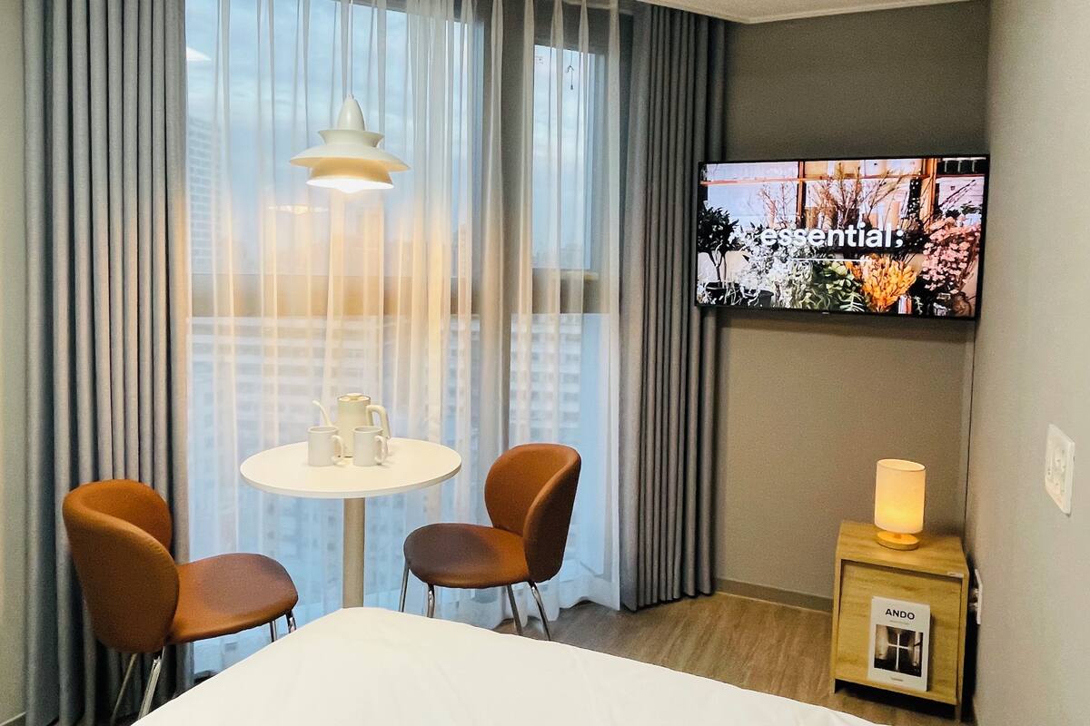 Habitación de hotel con televisión en la pared, una mesa redonda con sillas, una lámpara en una mesita de noche y una ventana con cortinas.