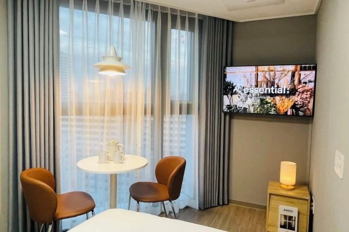Una habitación de hotel con un ventanal grande, un televisor de pantalla plana, una mesa redonda con dos sillas y una mesita de noche con lámpara.