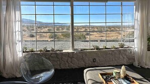 Une pièce dotée d’une grande fenêtre donnant sur un paysage désertique, avec un mur en pierre, un coin salon agrémenté de coussins et une petite table sur laquelle sont posés divers objets.