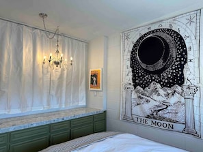 Quarto com um grande mural na parede intitulado 'A Lua', um candelabro e uma janela com cortinas.