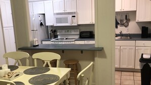 Una cocina con gabinetes blancos, un microondas, una estufa y un lavaplatos. Hay una mesa de comedor con sillas y un cartel encima que dice "BEACH".