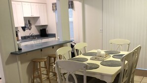 Un área de comedor con una mesa y sillas blancas, una cocina al fondo y un cartel que indica el camino a la playa.