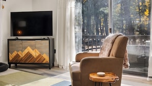 Een moderne woonkamer met een grote televisie, een bank en uitzicht op bomen door een groot raam.