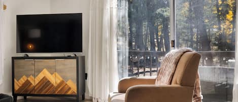 Una sala de estar moderna con un televisor grande, un sofá y vistas a árboles a través de un ventanal amplio.