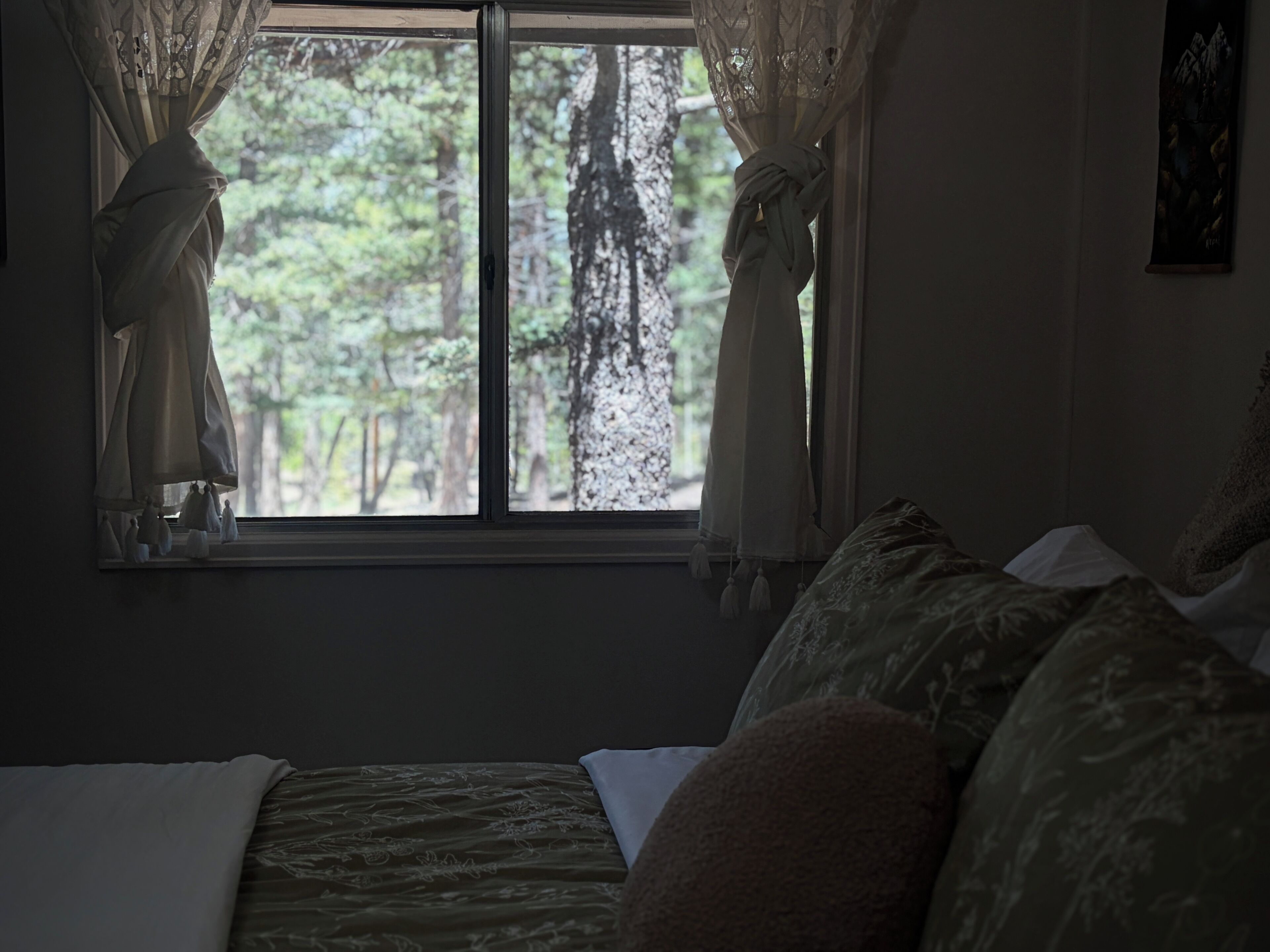 Ein Schlafzimmer mit einem Bett, einem Fenster mit Vorhängen und Blick auf Bäume.