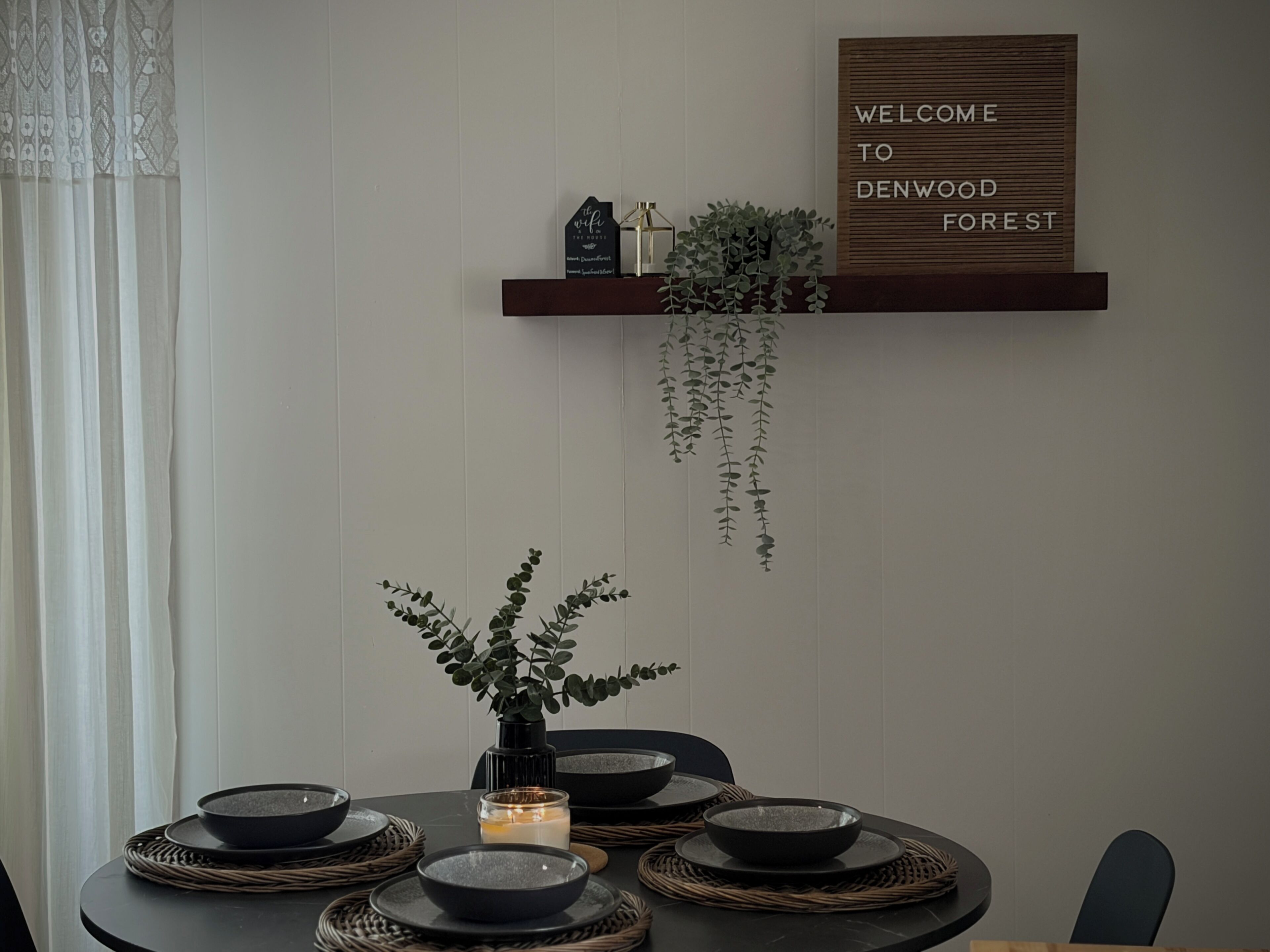A dining table with black bowls and a candle, a welcome sign, and a hanging plant.