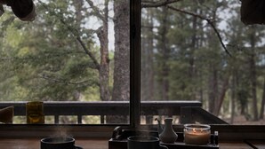 Een uitzicht op een bos, een houten tafel met twee mokken, een kaars en een theepot.