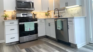 A modern kitchen with white cabinets, a stainless steel microwave, oven, and dishwasher. There are two potted plants and a wooden cutting board on the countertop.