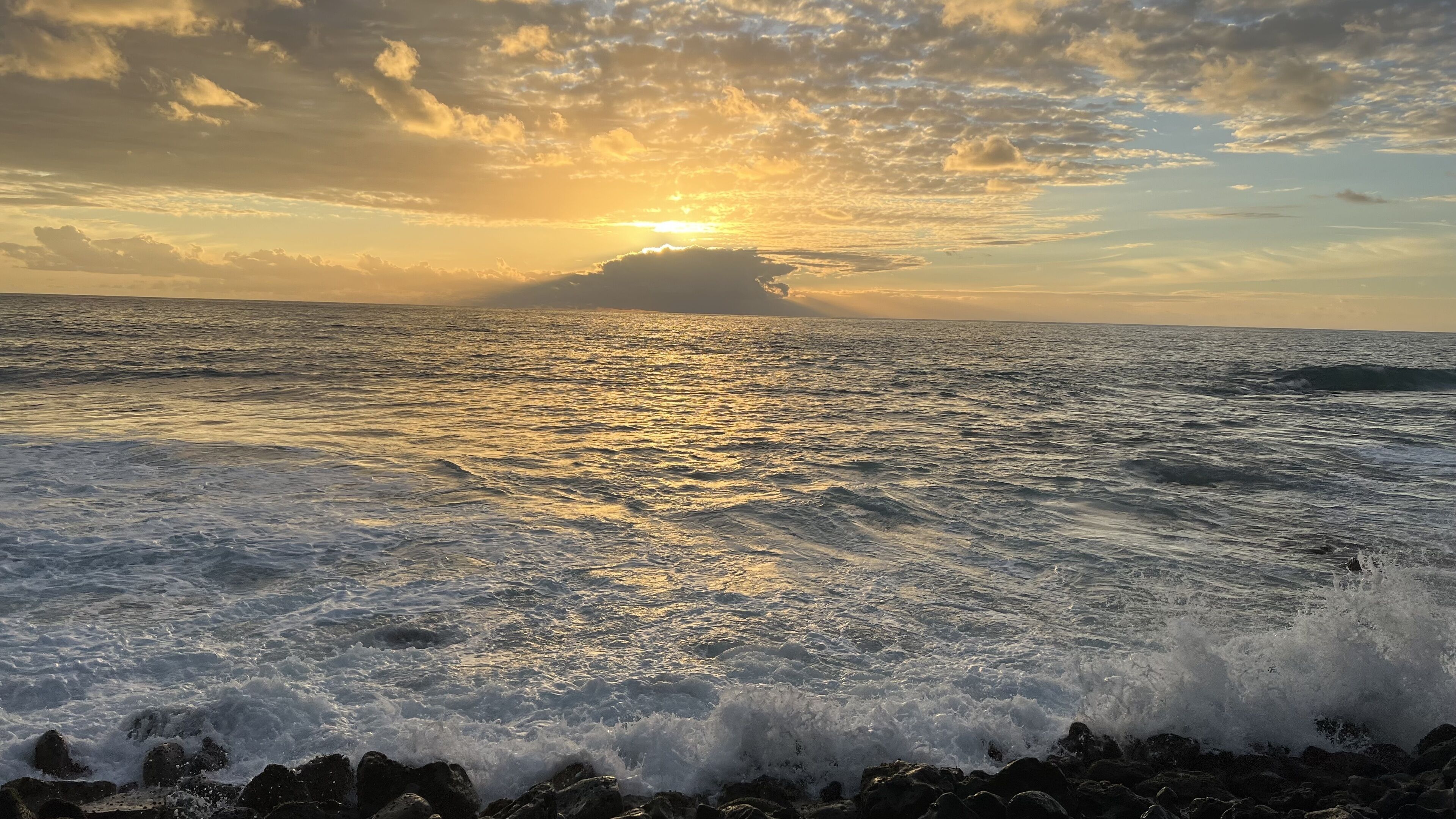 A coastal scene with waves crashing onto rocks, a clear sky, and a sun setting or rising near the horizon.