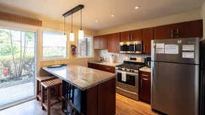 A modern kitchen with wooden cabinets, stainless steel appliances, a large island with bar stools, and a view through the sliding glass door. - Peek-a-boo Ocean View,1bd1bath AC Condo, closer to downtown Kailua village (Kailua-Kona)