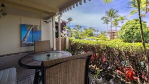 A covered outdoor seating area with a round table and chairs, a painting on the wall, and a view of palm trees and buildings in the background. - Peek-a-boo Ocean View,1bd1bath AC Condo, closer to downtown Kailua village (Kailua-Kona)