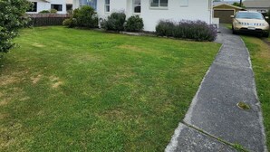 Un edificio residencial de dos plantas con jardín delantero, entrada para vehículos y un coche estacionado.