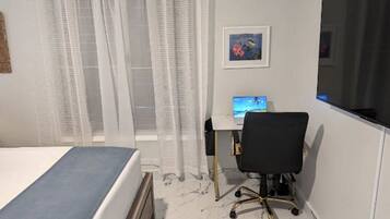 A bedroom with a bed, a desk with a computer, a chair, and a window with curtains.