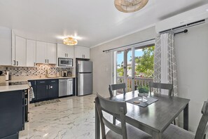 A modern kitchen with white cabinets, a stainless steel refrigerator, and a microwave. There is a dining table with chairs and a view of the outdoors through a window.