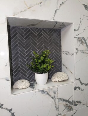 Une salle de bain avec un mur en marbre blanc, un pare-chocs en carreaux disposés en motif chevrons, un pot en blanc contenant des plantes vertes et deux pierres blanches à texture particulière.