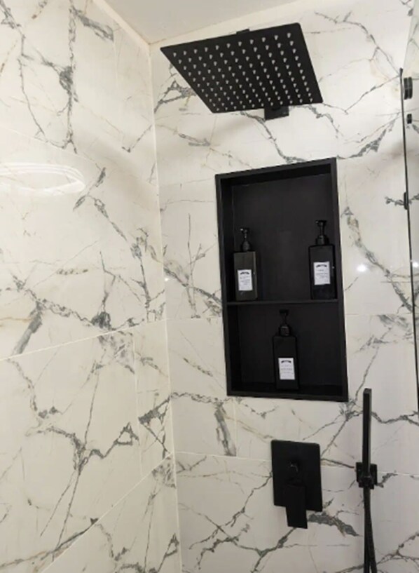 A bathroom with marble walls, a shower with a black ceiling panel, and three black shelves with bottles.