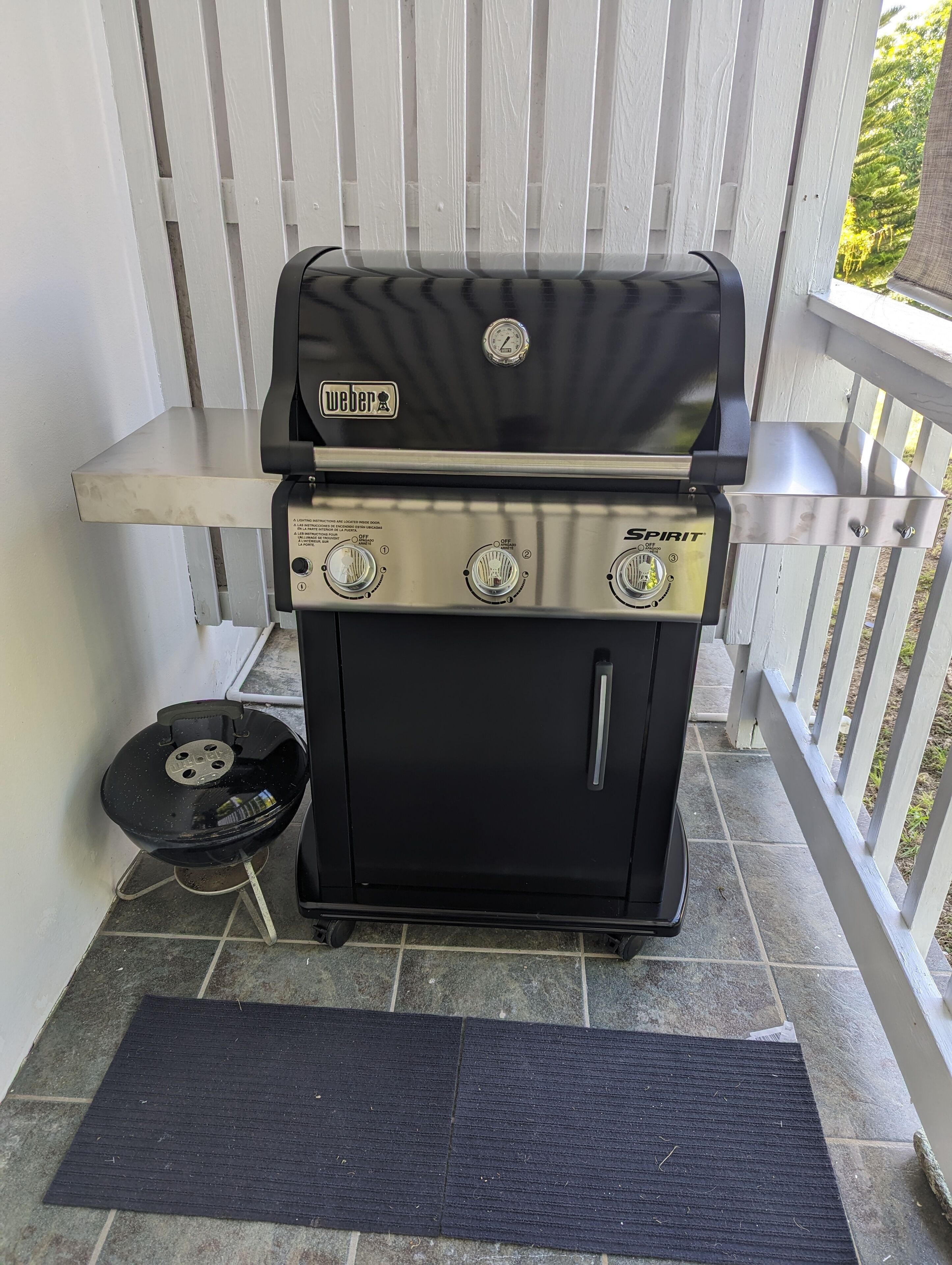 Een Weber Spirit draagbare barbecue op een terras met een witte balustrade en een zwarte mat.
