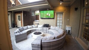 Un espacio de estar al aire libre con techo, que incluye un sofá curvo modular, un brasero y un televisor de pantalla plana montado en la pared.