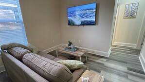 A living room with a large flat-screen TV mounted on the wall, a beige sofa, a small wooden coffee table, and a decorative wall piece. - Peachy Beach House - Unit A - Close to beach, game Room, Putting Green, & more! (Gulfport)