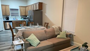 A living room with a brown sofa, a coffee table, a side table, and a dining area with a table set for four. - Peachy Beach House - Unit A - Close to beach, game Room, Putting Green, & more! (Gulfport)