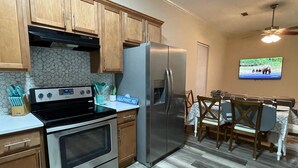 A kitchen with wooden cabinets, a stainless steel refrigerator, a stove, and a dining area with a table and chairs. - Peachy Beach House - Unit A - Close to beach, game Room, Putting Green, & more! (Gulfport)