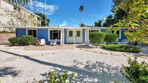 A two-story building with a blue exterior, a covered patio with white chairs, and a gravel driveway. - Renovated 2bed / 1bath North End of Anna Maria, 2min walk to the beach  (Anna Maria)