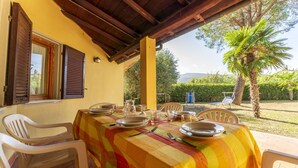 Un espacio comedor techado al aire libre con una mesa preparada para cuatro personas, rodeada de sillas de plástico blanco, bajo una pérgola de madera con vistas a un jardín y palmeras.