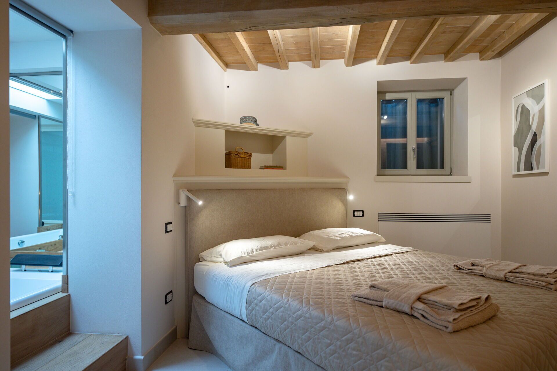 A bedroom with a bed, a window, a shelf with a handbag, and a painting on the wall.