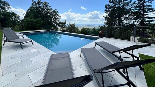 A rectangular swimming pool with a clear blue water surface, surrounded by a tiled deck. There are two lounge chairs on the deck, and the pool is bordered by a low fence.