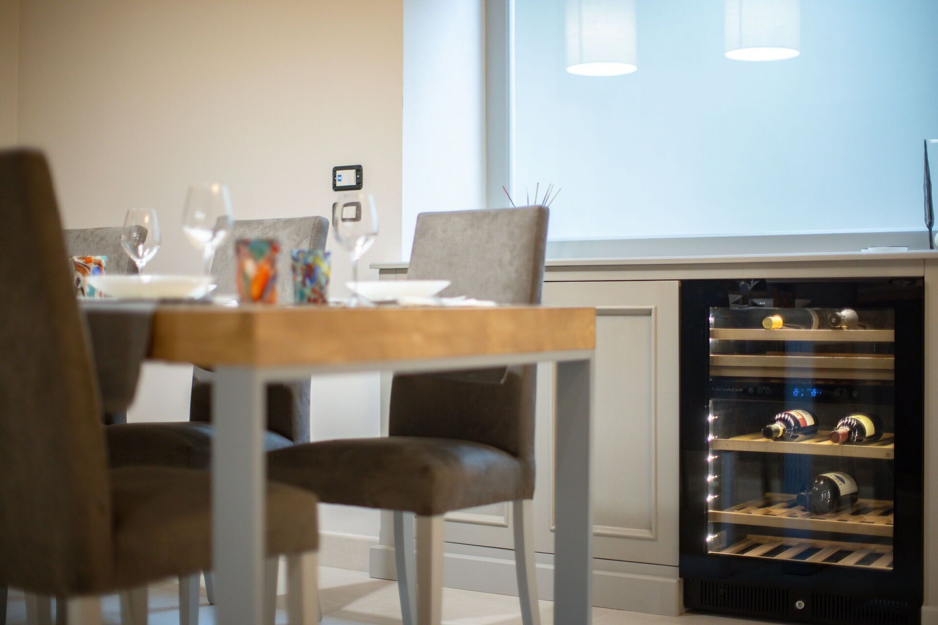 Un área de comedor con una mesa lista para dos personas, un enfriador de vino con botellas y una ventana con persianas.