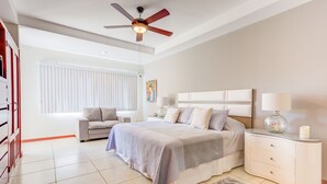 Un dormitorio con cama, mesitas de noche, ventilador de techo, ventana con persianas y una puerta.